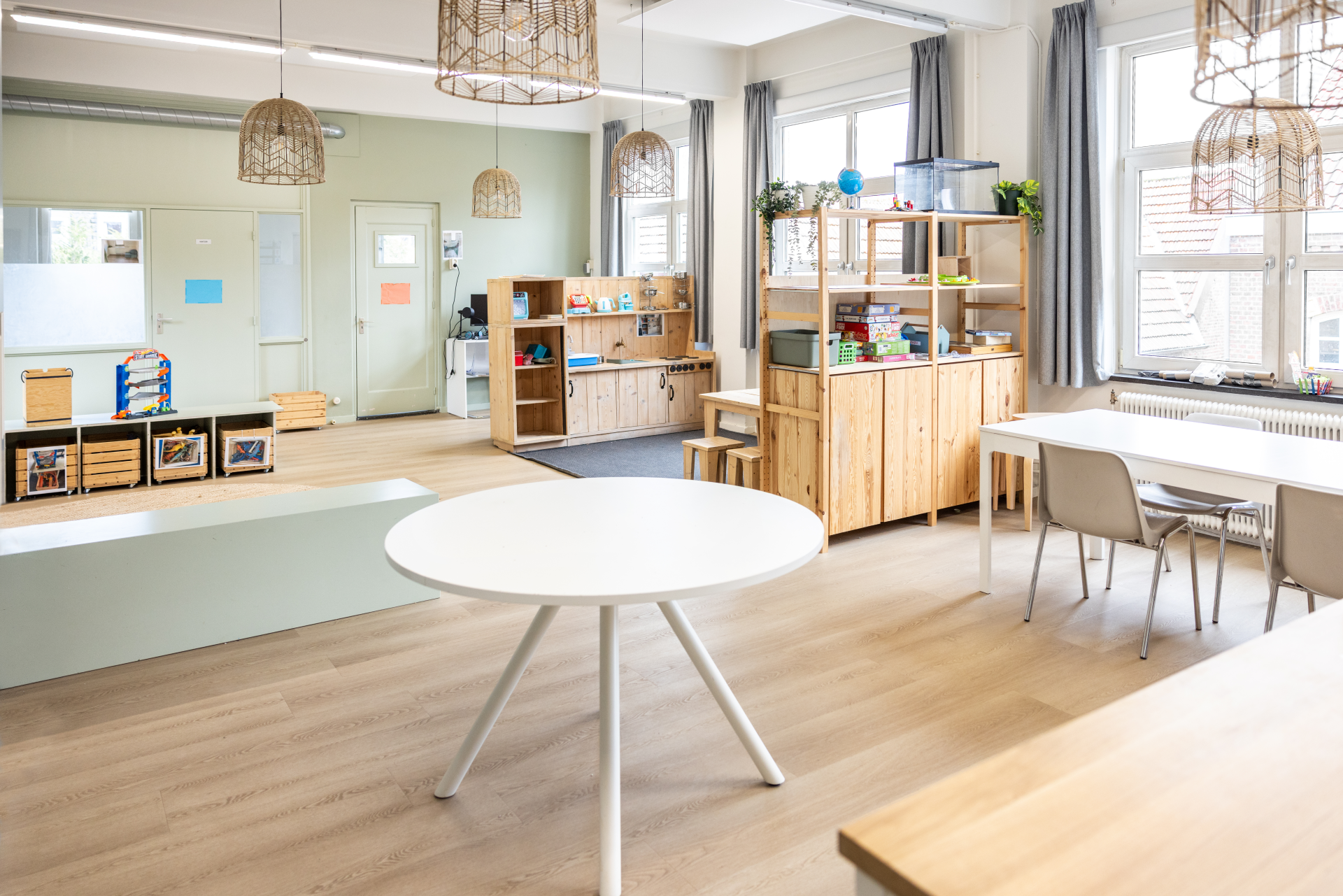 Lichte, ruime groepsruimte in een BSO met houten kasten, ronde tafels, speelhoeken en grote ramen die veel daglicht binnenlaten