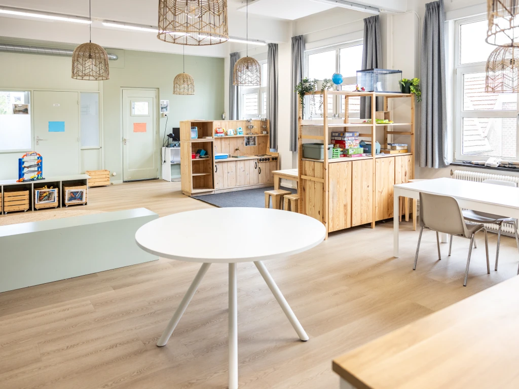 Lichte, ruime groepsruimte in een BSO met houten kasten, ronde tafels, speelhoeken en grote ramen die veel daglicht binnenlaten