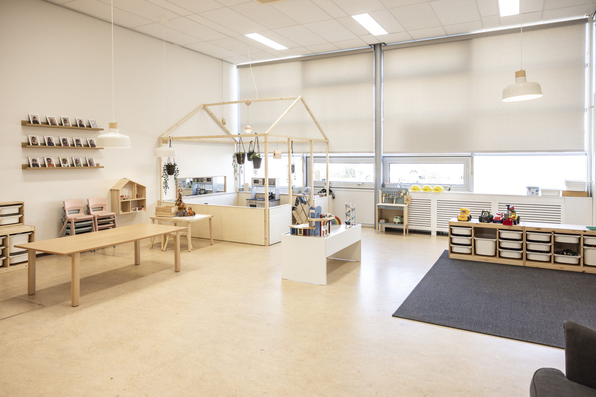 Lichte, ruim ingerichte kinderopvangruimte met houten meubels, speelhoeken, speelgoed en grote ramen.