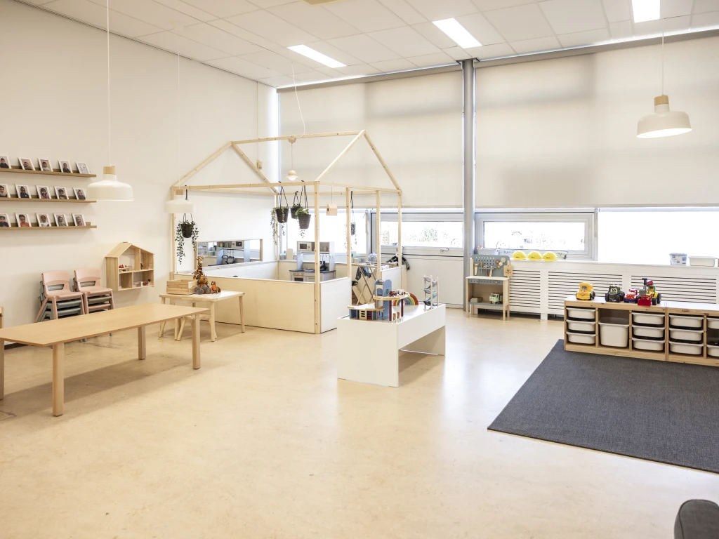 Lichte, ruim ingerichte kinderopvangruimte met houten meubels, speelhoeken, speelgoed en grote ramen.