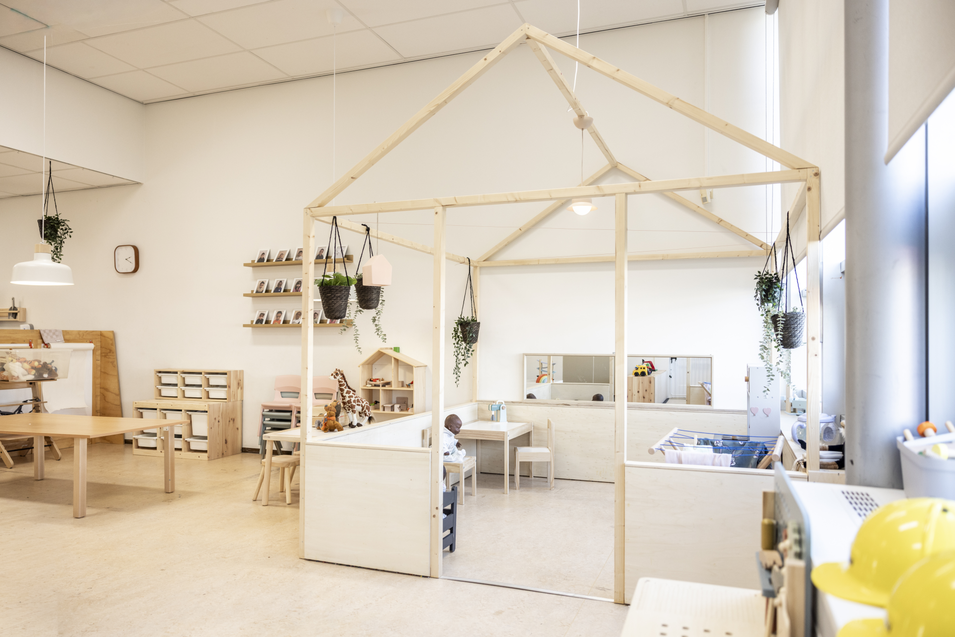 Lichte kinderopvangruimte met houten speelhoeken, een huisvormige constructie, hangplanten en verschillende speelmaterialen.