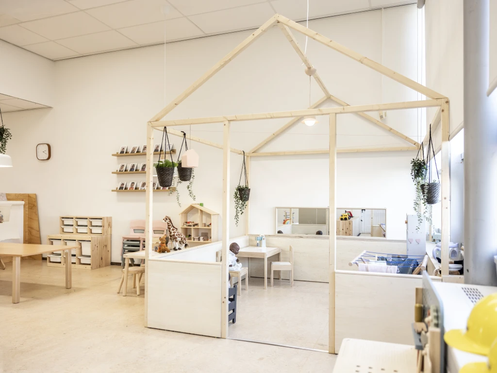 Lichte kinderopvangruimte met houten speelhoeken, een huisvormige constructie, hangplanten en verschillende speelmaterialen.
